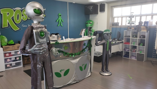 Two alien figures in a themed store with green decor and a reception desk, promoting extraterrestrial merchandise.