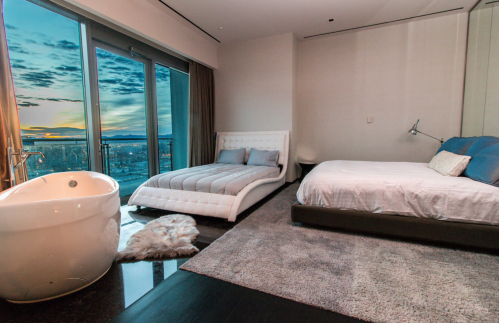 Modern bedroom with a large window, featuring a white bed, a gray bed, a freestanding bathtub, and a plush rug.