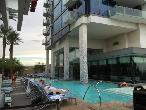 A modern hotel pool area with lounge chairs, palm trees, and a glass building in the background under a cloudy sky.