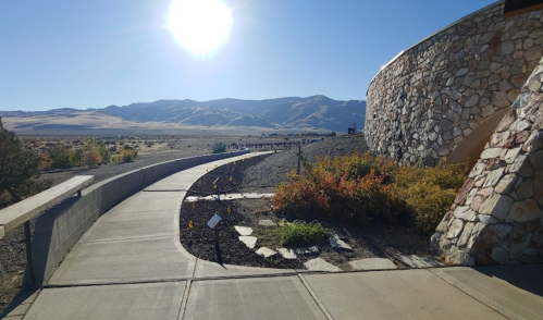 A sunlit pathway leads through a landscape with mountains in the background and a stone building on the right.