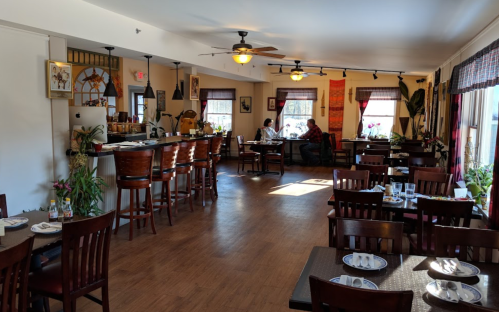 Cozy restaurant interior with wooden tables, bar seating, and patrons dining by the windows. Bright and inviting atmosphere.