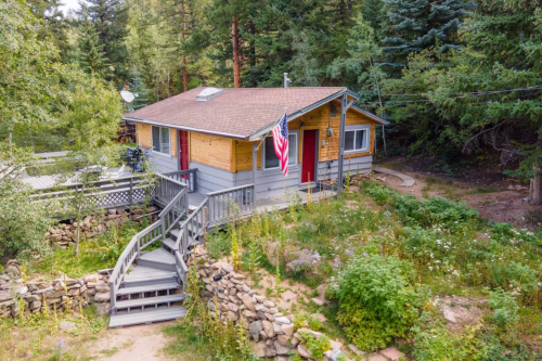 A cozy cabin surrounded by trees, featuring a wooden deck and an American flag, with a stone wall and garden.