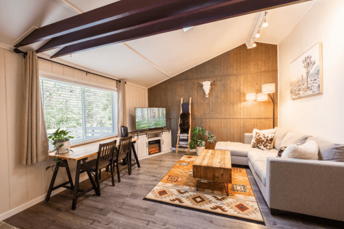 Cozy living room with a gray sofa, wooden dining table, TV, and rustic decor, featuring a warm, inviting atmosphere.