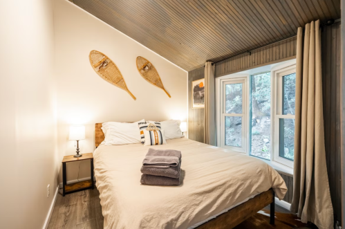 Cozy bedroom with a wooden bed, gray towels, and snowshoes on the wall, featuring a large window with natural light.