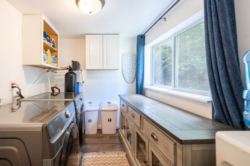 A bright laundry room featuring a washer, storage cabinets, a wooden table, and two recycling bins by a large window.