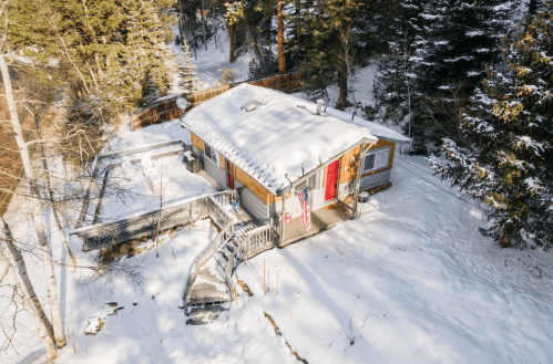 A cozy cabin surrounded by snow-covered trees, featuring a wooden deck and an American flag hanging by the entrance.