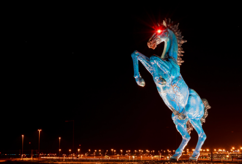 A large blue horse statue with glowing red eyes stands against a night sky, illuminated by surrounding lights.