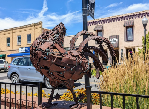 A metal sculpture of a rooster made from various tools and parts, set against a backdrop of shops and blue sky.