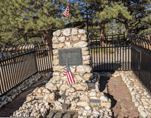 A stone monument surrounded by a black fence, featuring small American flags and trees in the background.