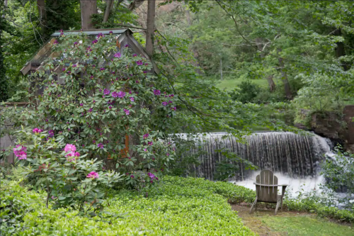A serene scene featuring a waterfall, lush greenery, and a chair beside blooming flowers.