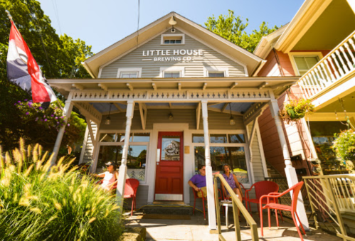A charming brewery with a red door, outdoor seating, and colorful flags, surrounded by greenery and sunny skies.