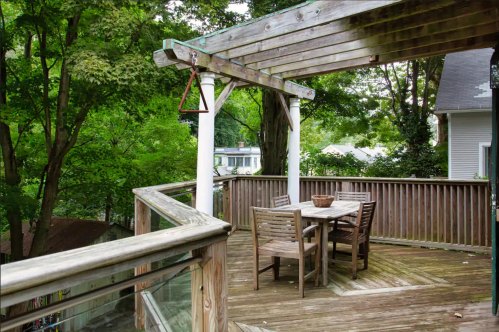 A wooden deck with a table and chairs, surrounded by lush green trees and a peaceful neighborhood view.