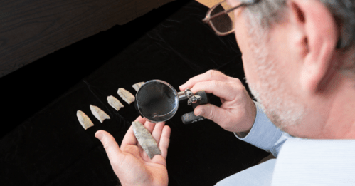 A person examines a stone artifact with a magnifying glass, with several similar artifacts displayed in front.
