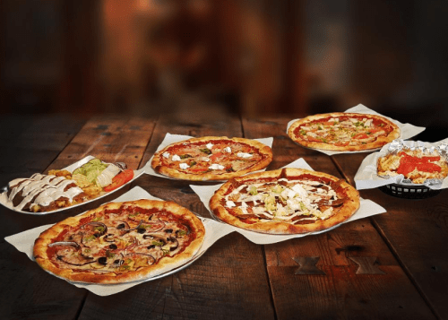 A variety of pizzas and dishes displayed on wooden tables, featuring toppings like vegetables, meats, and cheese.