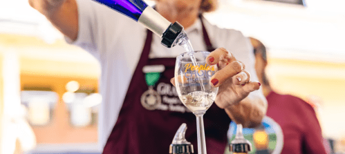 A person in an apron pours a drink into a glass at an event, with bottles and another person in the background.