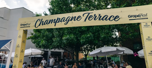 A yellow archway sign reading "Champagne Terrace" at an outdoor event with tables and umbrellas.