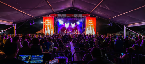 A lively outdoor concert scene with a large crowd enjoying a performance under a tent, colorful lights illuminating the stage.