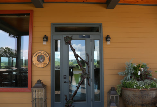Entrance to a tasting room with a decorative door and sign, surrounded by plants and lanterns. Scenic view in the background.