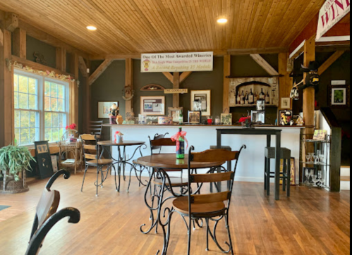 Cozy winery interior with wooden beams, tables, and a bar displaying awards and photos. Red flowers on tables.