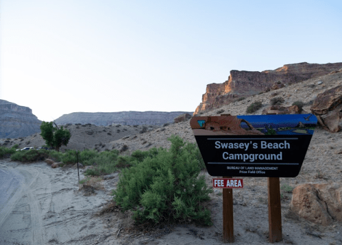 Sign for Swasey's Beach Campground in a desert landscape, with rocky cliffs in the background and a sandy path.