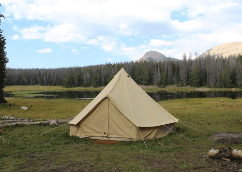 A beige tent set in a grassy area near a calm lake, surrounded by trees and mountains under a partly cloudy sky.