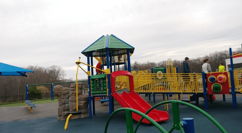 Colorful playground equipment with slides and climbing structures, set against a cloudy sky and trees in the background.