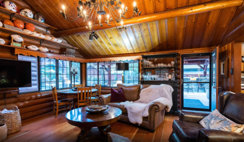 Cozy cabin interior with wooden walls, a chandelier, comfortable seating, and a view of the outdoors through large windows.