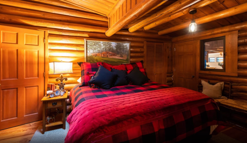 Cozy log cabin bedroom with a red and black plaid bedspread, wooden walls, and warm lighting.