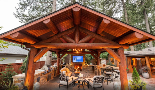 Cozy outdoor patio with a wooden gazebo, seating area, fireplace, and a TV, surrounded by trees.