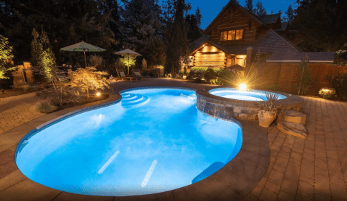 A beautifully lit pool and spa area at night, surrounded by trees and a cozy wooden house in the background.