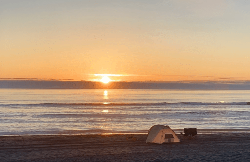 A serene beach at sunset, with gentle waves and a tent set up on the sand.