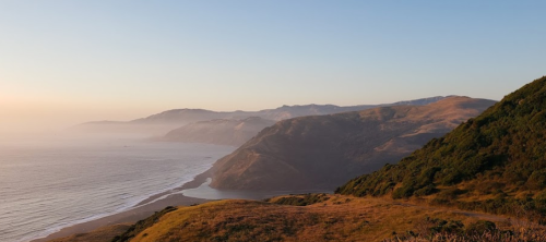 A serene coastal landscape at sunset, featuring rolling hills and a calm ocean along the shoreline.