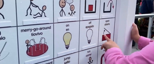 A child points at a communication board with symbols for activities and concepts, including a merry-go-round and a light bulb.