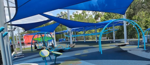 Colorful playground with blue shade sails, swings, and various play structures on a rubber surface surrounded by greenery.