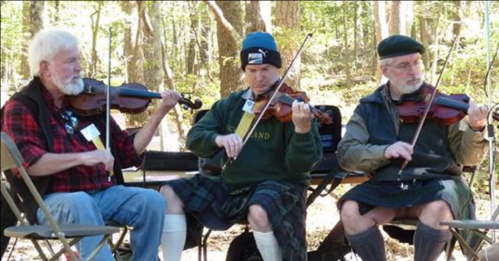 Three men playing violins outdoors, dressed in casual attire and kilts, seated on folding chairs in a wooded area.