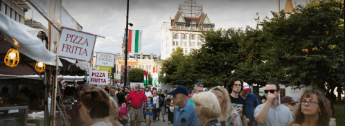 Crowd of people walking through a street fair with food stalls and festive decorations in the background.