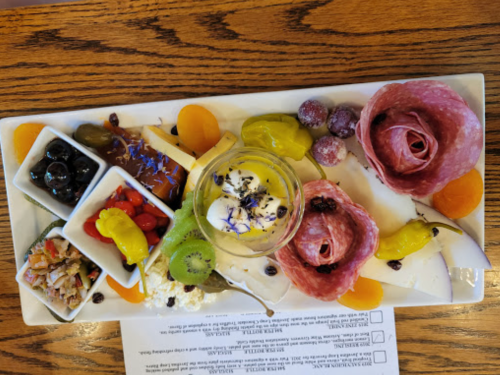 A colorful platter featuring assorted fruits, meats, olives, and a small dessert, arranged artistically on a white plate.