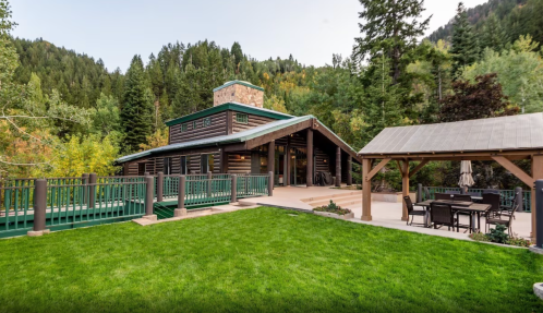 A modern log cabin surrounded by trees, featuring a spacious lawn and a patio with outdoor seating.