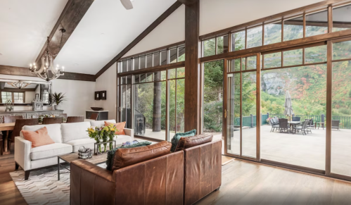 Cozy living room with large windows, wooden beams, and a view of a patio and nature outside.
