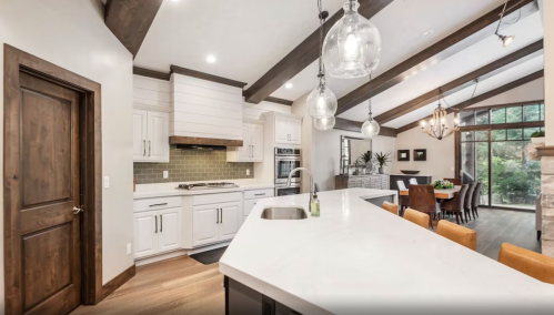 Modern kitchen with white cabinets, a large island, pendant lights, and a dining area with a view of greenery outside.