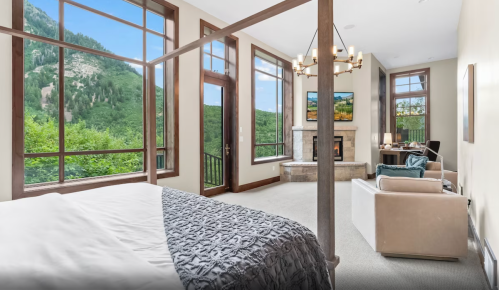 Spacious bedroom with large windows overlooking mountains, featuring a cozy seating area and a fireplace.