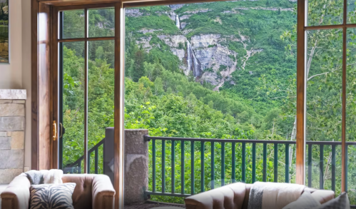 A cozy living room with large windows showcasing a lush green mountain landscape and a waterfall in the distance.