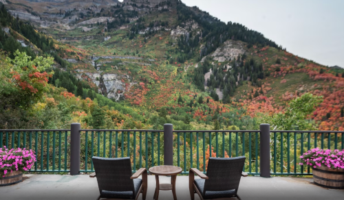 A scenic view from a balcony overlooking a colorful autumn landscape with mountains and trees. Two chairs and a table are visible.