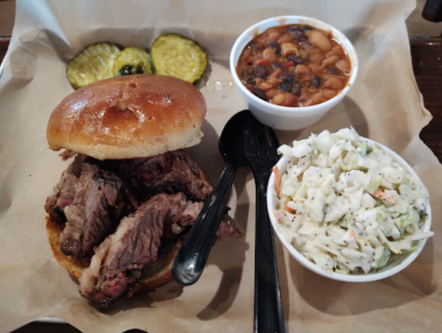 A plate featuring a beef sandwich, pickles, coleslaw, and a bowl of baked beans.