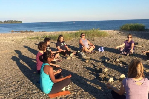 A group of people meditating in a circle on a beach, with the ocean and a clear sky in the background.