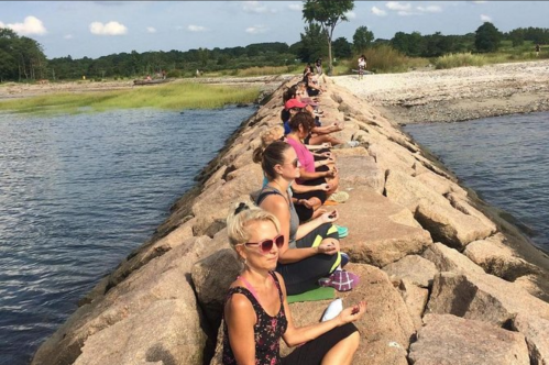 A group of people meditating on a rocky jetty by a calm water body, surrounded by greenery and blue skies.