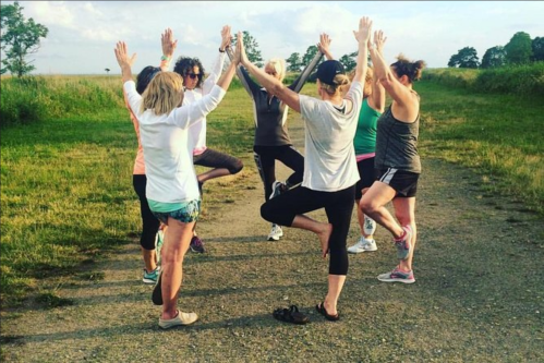 A group of women practicing yoga outdoors, balancing on one leg with arms raised, surrounded by greenery.