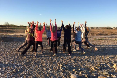 A group of women stands in a circle outdoors, raising their arms towards the sky against a sunset backdrop.
