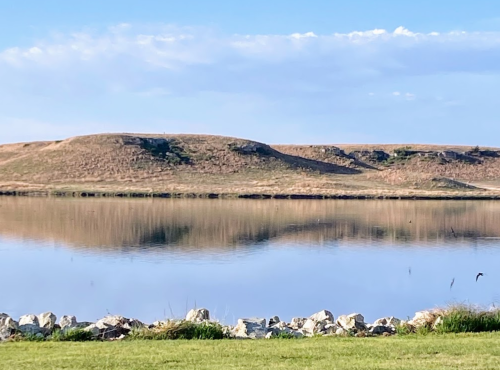 A calm lake reflects grassy hills under a clear blue sky, with a rocky shoreline in the foreground.