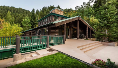 A modern log cabin surrounded by trees, featuring a green roof and a welcoming entrance with steps and a ramp.
